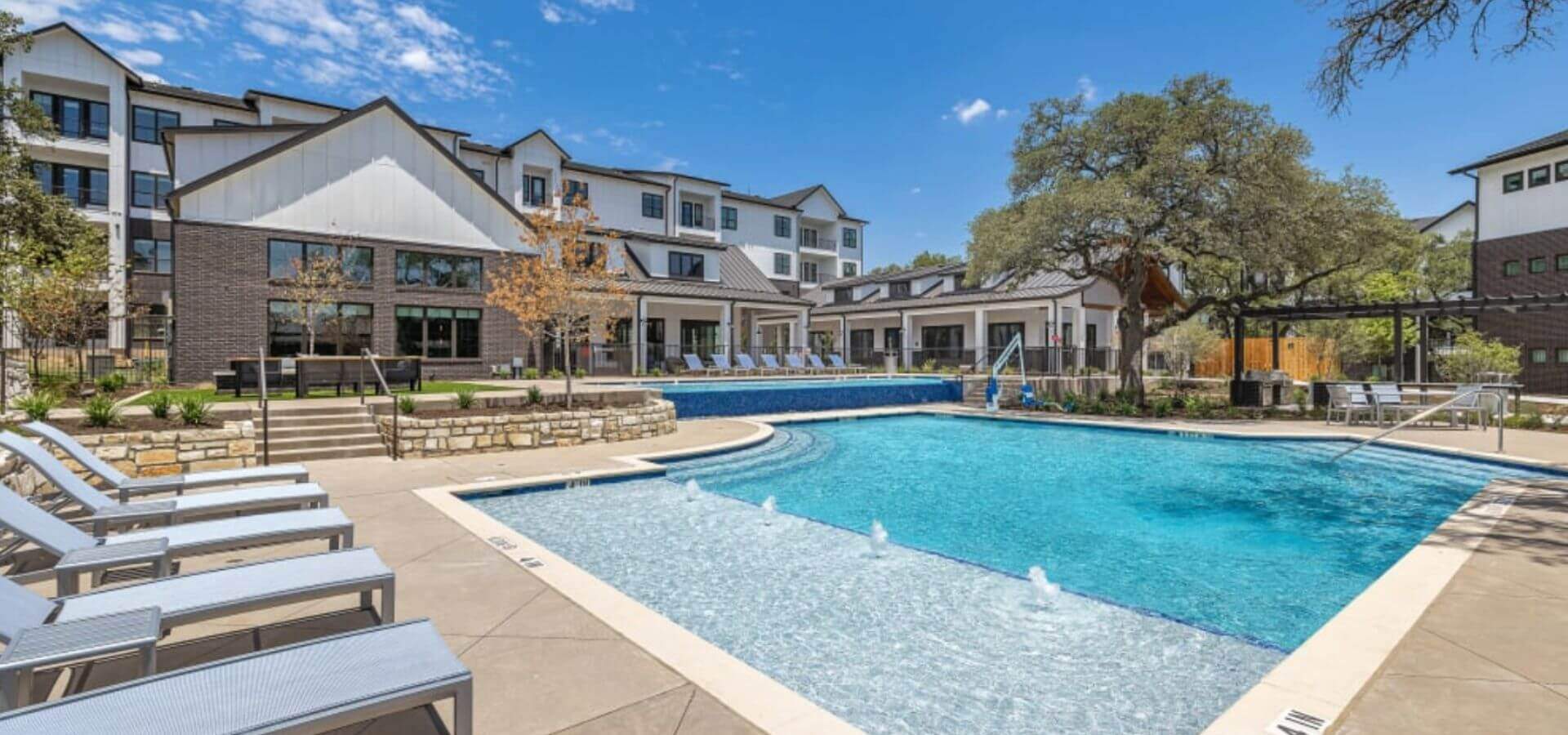 The Avery by Compass Furnished Apartments — Outdoor resort-style pool with lounge seating, shallow wading area, and view of modern residential buildings in the background.