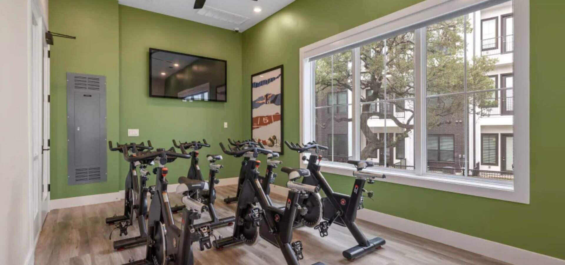 The Avery by Compass Furnished Apartments — Spin studio featuring multiple stationary bikes, large windows with natural light, a green accent wall, and a mounted TV for interactive workouts.