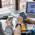 Two people reviewing spreadsheets, calculating expenses on a smartphone, and analyzing graphs on a laptop—depicting the process of managing taxes and regulatory compliance for corporate housing.
