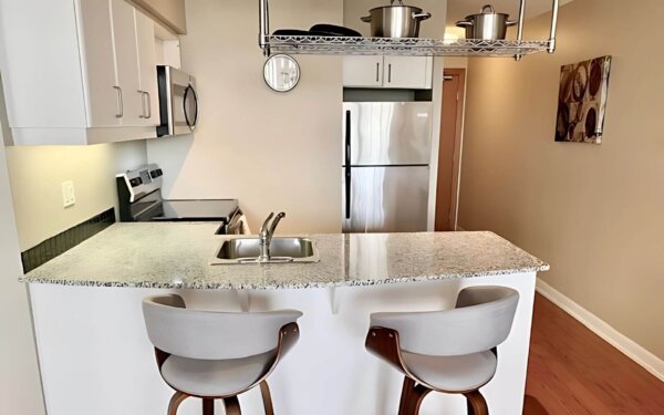Contemporary kitchen at MAC Garden by Compass Furnished Apartments, featuring granite countertops, stainless steel appliances, overhead pot rack, and a breakfast bar with modern swivel stools.