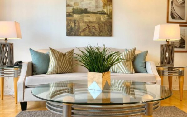 Stylish living room at MAC Garden by Compass Furnished Apartments, featuring a cream sofa with striped and teal pillows, glass coffee table, and decorative indoor plant.
