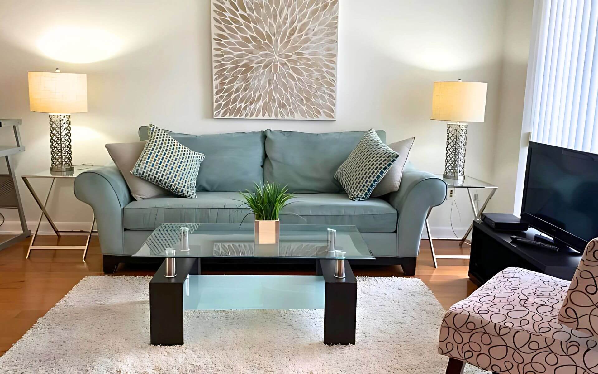 Stylish living room at MAC Garden by Compass Furnished Apartments, featuring a soft teal sofa with patterned pillows, glass-top coffee table, and contemporary lighting.