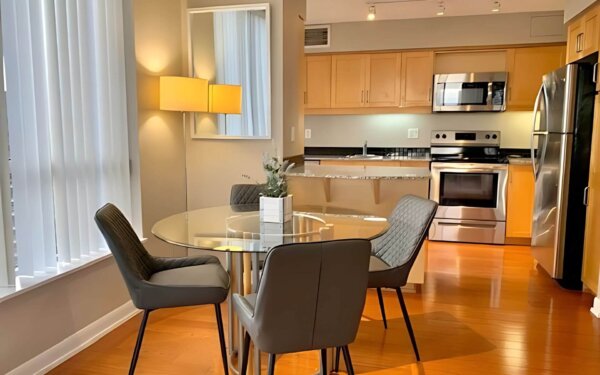 Stylish kitchen and dining space at MAC Garden by Compass Furnished Apartments, featuring a round glass dining table, modern gray chairs, stainless steel appliances, and warm wood cabinetry.