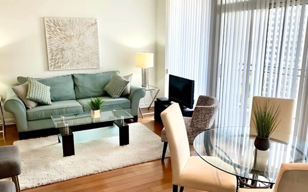 Bright and stylish living room at MAC Garden by Compass Furnished Apartments, featuring a teal sofa, glass coffee table, patterned accent chair, and round glass dining table by floor-to-ceiling windows.