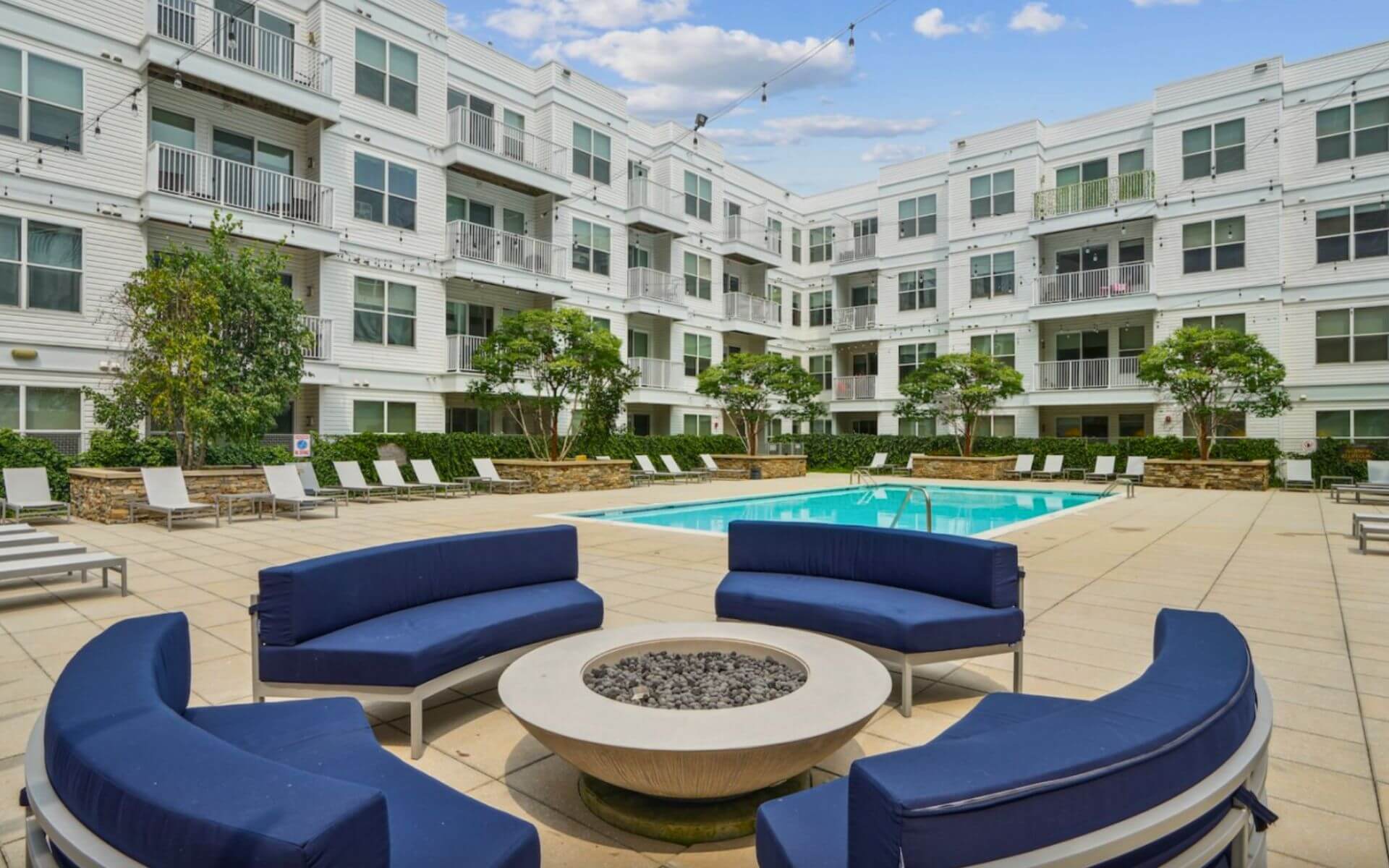Outdoor courtyard at 75 Tresser by Compass Furnished Apartments featuring a pool, firepit lounge, and modern seating surrounded by white apartment buildings.