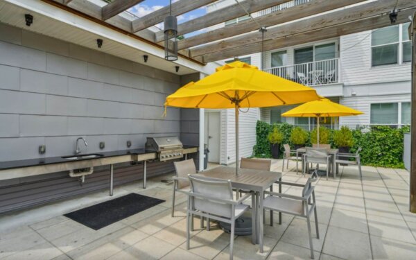 Outdoor grilling and dining area at 75 Tresser by Compass Furnished Apartments featuring stainless steel BBQ stations, yellow umbrellas, and modern patio seating.
