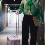 Business traveler walking through a hotel hallway with a pink suitcase and a laptop in hand.
