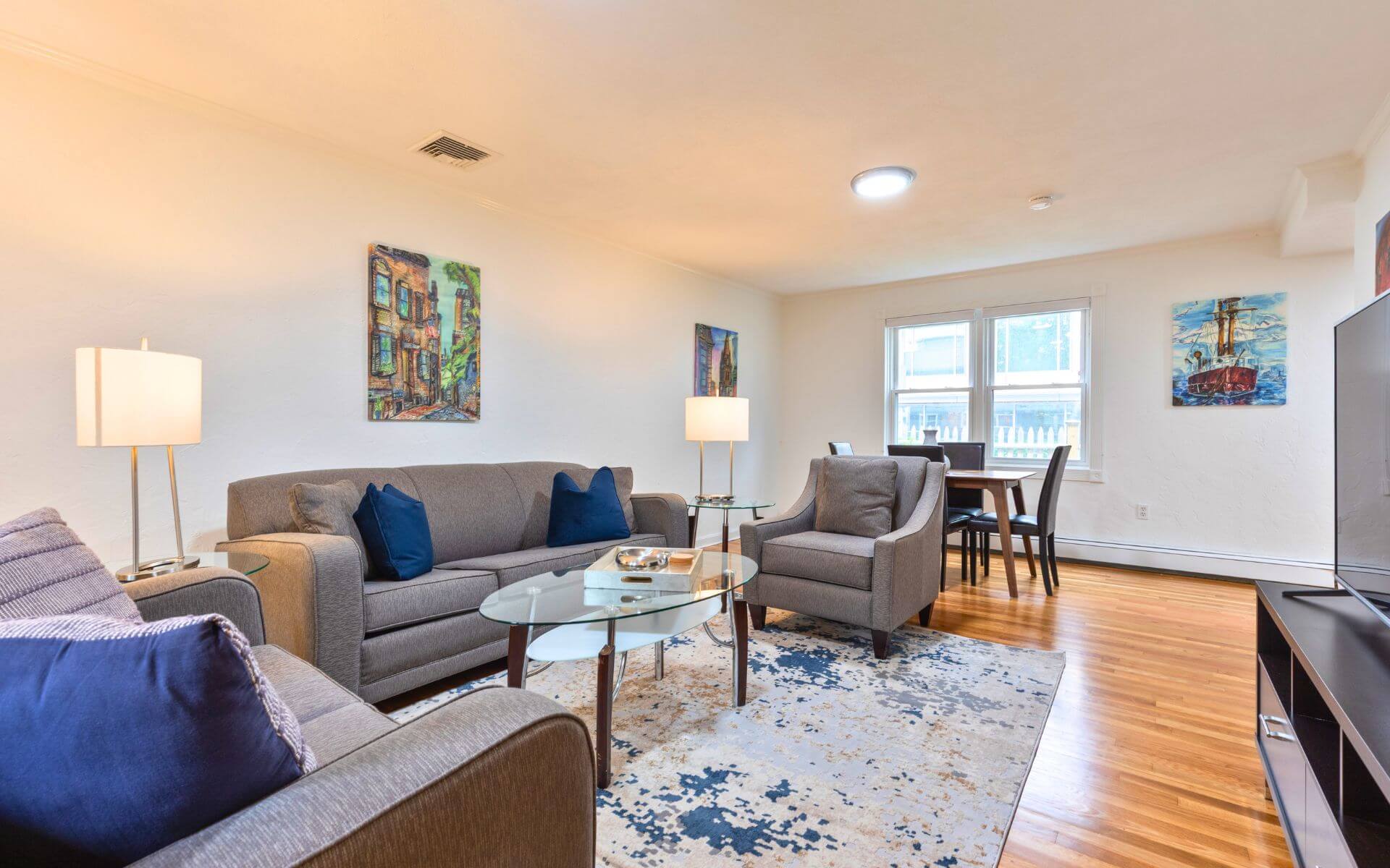 Bright and modern living room with grey sofas, accent chairs, glass coffee table, and dining area at Compass Furnished Apartments, 235 Cardinal Medeiros.
