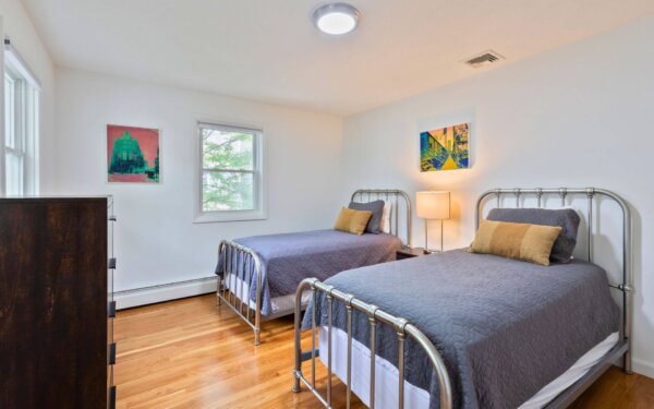 Twin bedroom with metal bed frames, warm lighting, hardwood floors, and modern art at Compass Furnished Apartments, 235 Cardinal Medeiros.