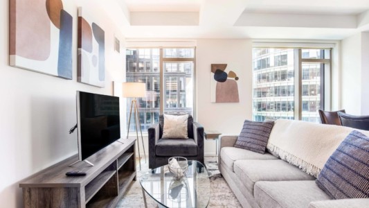Open-plan living area in a furnished temporary apartment, featuring a plush sofa set and a large window with city skyline views